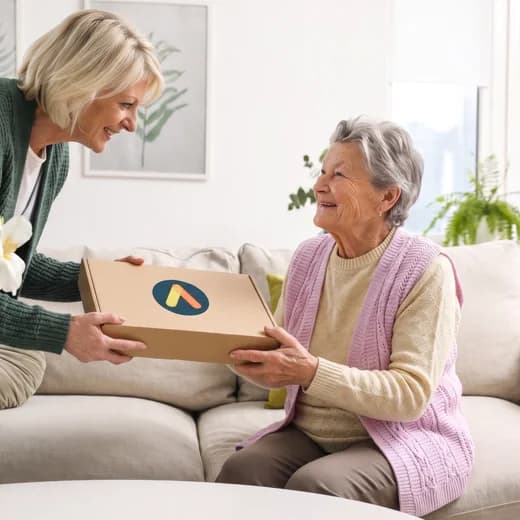Eine jüngere Frau überreicht einer älteren Frau das Amara-Paket auf dem Sofa